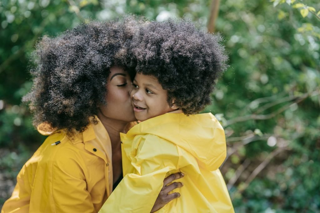 Mother lovingly kisses her daughter wearing yellow raincoats in a lush green outdoor setting.